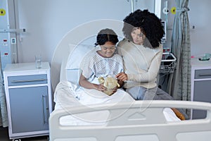 Mixed race mother comforting her sick daughter holding teddy bear in hospital bed