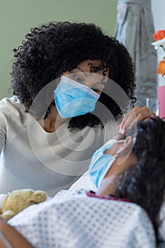 Mixed race mother and sick daughter in face masks in hospital, girl holding teddy bear