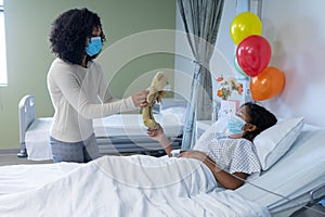 Mixed race mother and sick daughter in face masks in hospital, playing with teddy bear