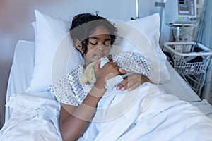 Mixed race sick girl asleep in hospital bed wearing fingertip pulse oximeter and holding teddy bear