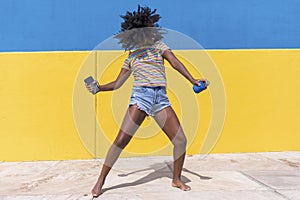 Mixed race smiling black woman portrait with big afro curly hair against blue and yellow wall dancing while holding a smartphone