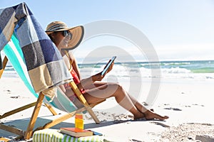 Mixed race smiling woman on beach holiday sitting in deckchair using tablet