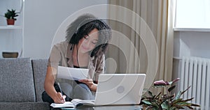 Mixed race student girl sits in room at laptop with headphones, reads documents, answers questions in online video