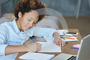 Mixed race teen schoolgirl looking focused while drawing on paper with colorful markers, spending time at home during