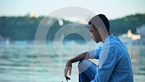 Mixed-race teenager throwing pebbles into river trying to relax, peaceful place