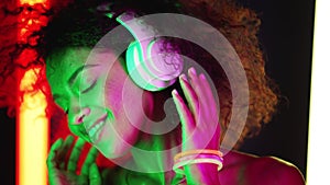 Mixed race woman with afro hairstyle dancing in wireless headphones in room with colorful neon lamps. Party girl moving
