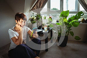 Mixed race woman in casual home clothes serfing in her smart phone sitting near coffee table with closed laptop and many