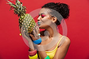 Mixed-race woman with fashion makeup in yellow shirt having detox kissing fresh ripe pineapple holding in hands isolated, over re