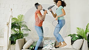 Mixed race young funny girls dance singing with hairdryer and comb jumping on sofa. Sisters having fun leisure in living