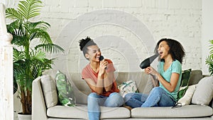 Mixed race young funny girls dance singing with hairdryer and comb sitting on sofa. Sisters having fun leisure in living