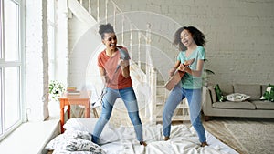 Mixed race young funny girls dance singing with hairdryer and playing acoustic guitar on a bed. Sisters having fun