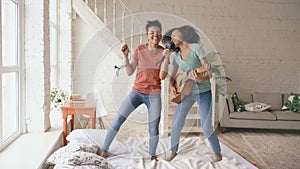 Mixed race young funny girls dance singing with hairdryer and playing acoustic guitar on a bed. Sisters having fun