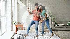 Mixed race young funny girls dance singing with hairdryer and playing acoustic guitar on a bed. Sisters having fun