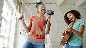 Mixed race young funny girls dance singing with hairdryer and playing acoustic guitar on a bed. Sisters having fun