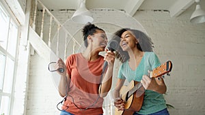 Mixed race young funny girls dance singing with hairdryer and playing acoustic guitar on a bed. Sisters having fun