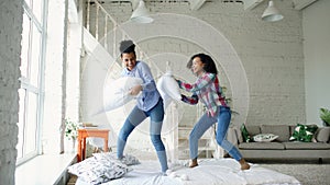 Mixed race young pretty girls jumping on bed and fight pillows having fun at home