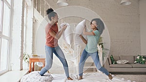 Mixed race young pretty girls jumping on bed and fight pillows having fun at home