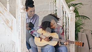Mixed race young woman sitting on stairs teaching her teenage sister to play acoustic guitar at home