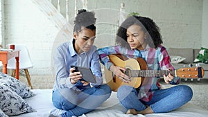 Mixed race young woman with tablet computer sitting on bed teaching her teenage sister to play acoustic guitar at home