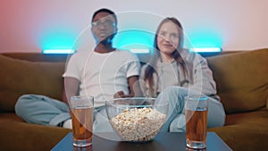 Mixed young couple watching fun movie while sitting on sofa at table with drinks and popcorn