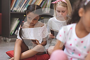 Mixte ethnicity schoolgirls holding digital tablet and sitting on cushions in a library