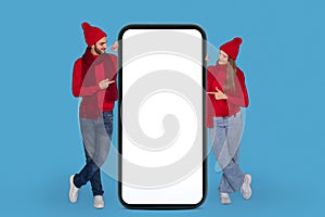 Mobile Ad. Smiling Couple Wearing Winter Hats Pointing At Huge Blank Smartphone