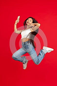 always on mobile. Full length of pretty young woman taking phone while jumping against red studio background.