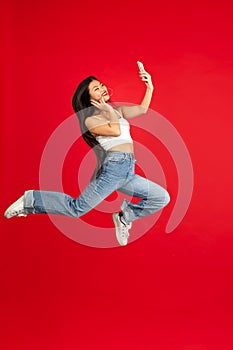always on mobile. Full length of pretty young woman taking phone while jumping against red studio background.