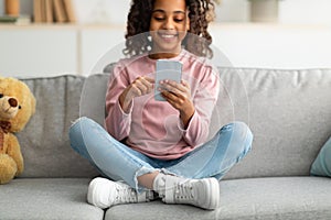 Mobile gaming concept. Happy african american teen girl using smartphone, surfing internet, sitting on sofa, closeup