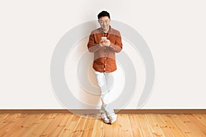 Mobile lesiure concept. Asian middle aged man using cellphone, surfing internet online standing against white wall