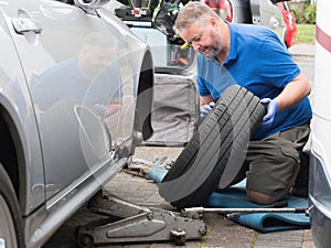 A mobile mechanic has jacked up a sports car and inspects the tyre and wheel which he has removed.The man kneels in shorts