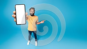 Mobile offer. Indian man showing phone with blank screen and pointing finger aside at free space on blue background