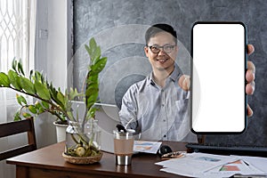 Mobile Offer. Smiling Young Man Showing Big Blank Smartphone While Sitting At Desk In Office, Happy Millennial Businessman