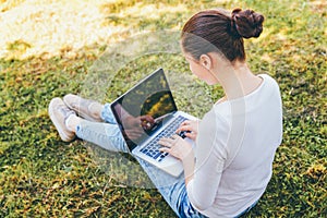 Mobile Office. Freelance business concept. Young woman sitting on green grass lawn in city park working on laptop pc computer.