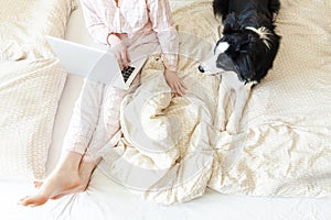 Mobile Office at home. Young woman in pajamas sitting on bed with pet dog working using on laptop pc computer at home. Lifestyle