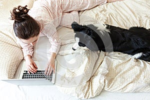 Mobile Office at home. Young woman in pajamas sitting on bed with pet dog working using on laptop pc computer at home. Lifestyle
