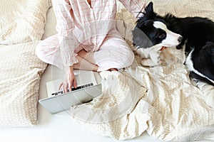 Mobile Office at home. Young woman in pajamas sitting on bed with pet dog working using on laptop pc computer at home. Lifestyle