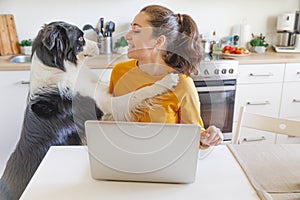 Mobile Office at home. Young woman with pet dog sitting in kitchen at home working using on laptop computer pc