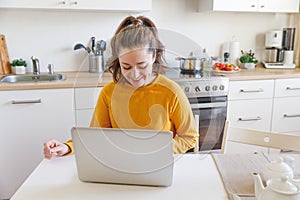 Mobile Office at home. Young woman sitting in kitchen at home working using on laptop computer. Lifestyle girl studying or working