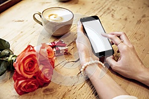 Mobile phone in beautiful woman hands. Lady writing message. Red roses flowers and present box behind on wooden table. St. Valenti