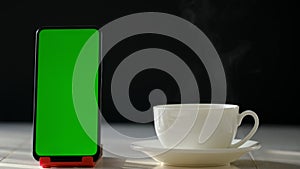 Mobile phone with blank green screen, front view, isolated on black background. There is a cup of hot coffee nearby