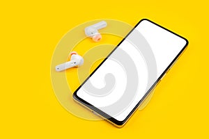 Mobile phone with empty screen and earphones on yellow surface. Wireless earphones, mobile phone on yellow background