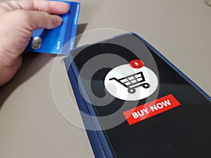 A mobile phone showing Buy Now shopping cart and off focus of a person holding a credit card