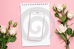 Mobile phone with a white notebook and roses flowers on pink background