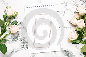 Mobile phone with a white and pink notebook and roses flowers on a marble background