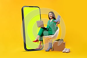 Mobile shopping concept. Lady sitting near giant cellphone and holding new shoes, buying online over yellow background