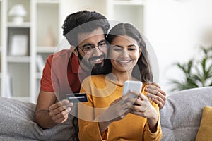 Mobile Shopping. Happy Indian Couple Using Smartphone And Credit Card At Home