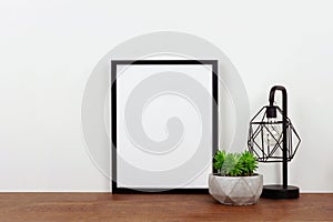 Mock up black frame against white wall with plant and lamp on a wood shelf