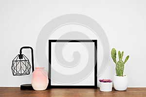 Mock up black square frame with home decor and potted plants on a wood shelf against a white wall