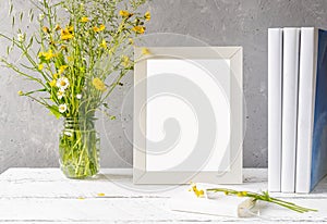 Mock up creation. White frame on white wooden table with grey concrete background, books, notebook and wild flowers